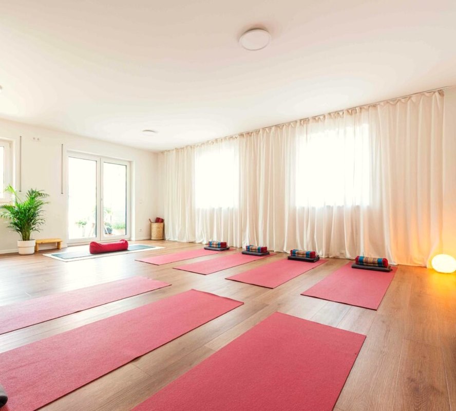 Unser „Großer“: der helle Yogaraum mit Vorhängen, roten Matten und Grünpflanze
